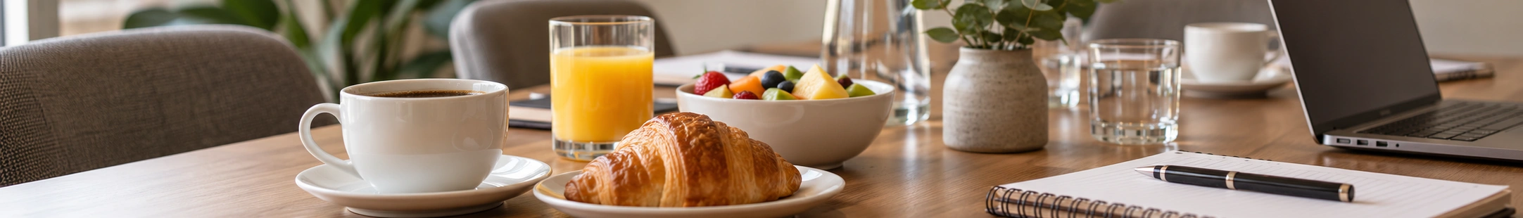Frühstück am Besprechungstisch mit Kaffee, Croissant, Obst, Orangensaft und Notizbuch